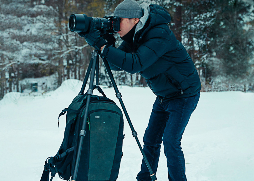 Person in winter clothing takes a photo with a camera on a tripod in a snowy landscape.
