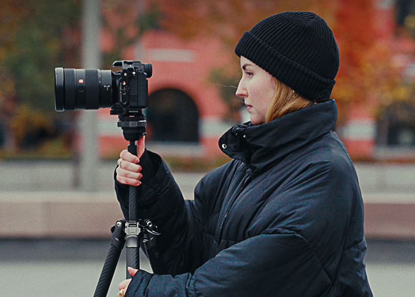 Person in a black coat and hat adjusts a camera on a tripod outdoors, with blurred autumn foliage and buildings in the background.