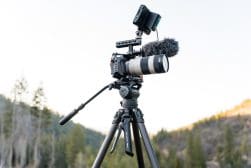 Professional video camera with a mounted microphone and telephoto lens on a tripod, set up outdoors with a forested backdrop.