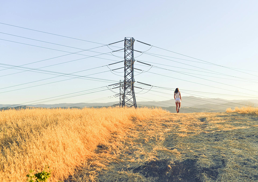 A person walks near a field with tall dry grass, next to an electricity pylon, under a clear sky.