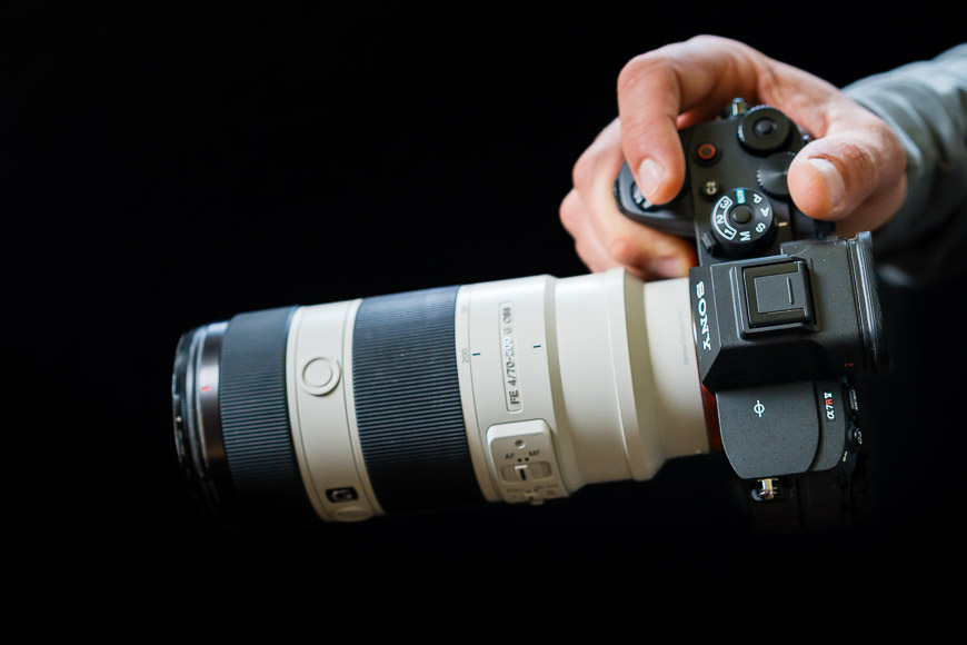 A hand holding a Sony camera with a large telephoto lens against a dark background.