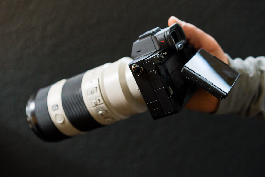 A person holds a camera with a large telephoto lens and an extended LCD screen, angled slightly upwards, against a dark background.