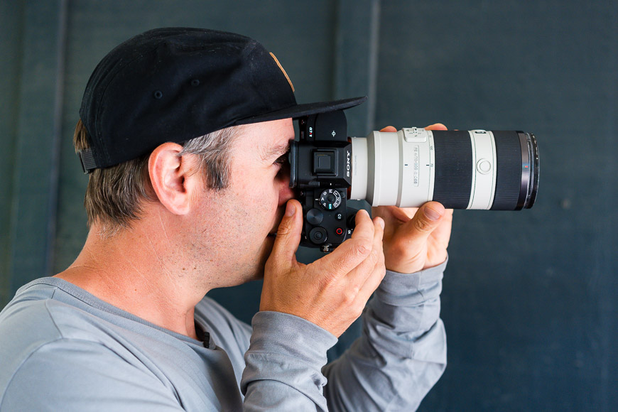 Person wearing a cap and gray shirt using a camera with a large zoom lens.