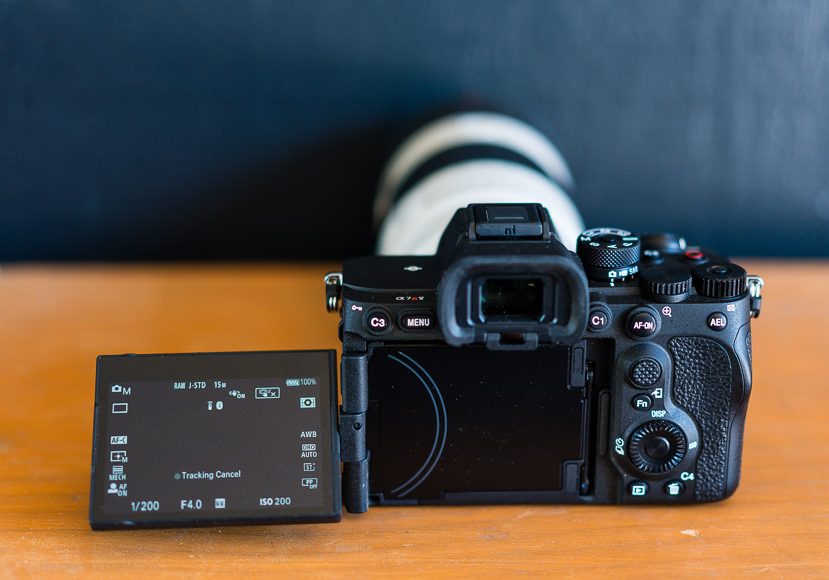 A digital camera with a large lens is on a wooden surface. The screen is flipped out, displaying various camera settings. A digital camera with a large lens is on a wooden surface. The screen is flipped out, displaying various camera settings.