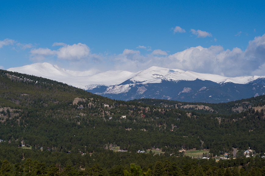 Mountain landscape with snow-capped peaks in the background under a blue sky, and a forested area with scattered buildings in the foreground.
