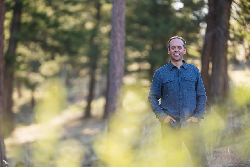 A man stands outdoors in a forested area, wearing a blue shirt and smiling.