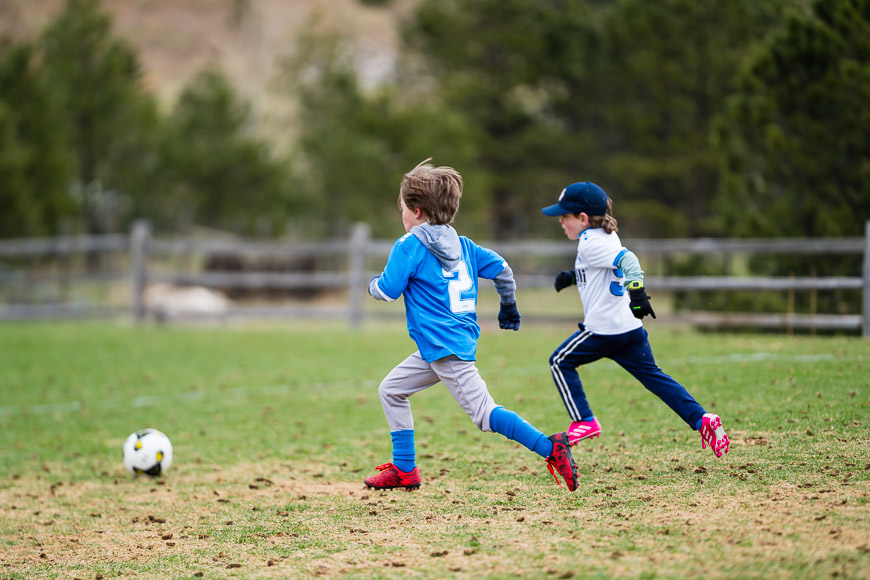 Two children wearing sports attire running towards a soccer ball on a grassy field with a wooden fence and trees in the background.