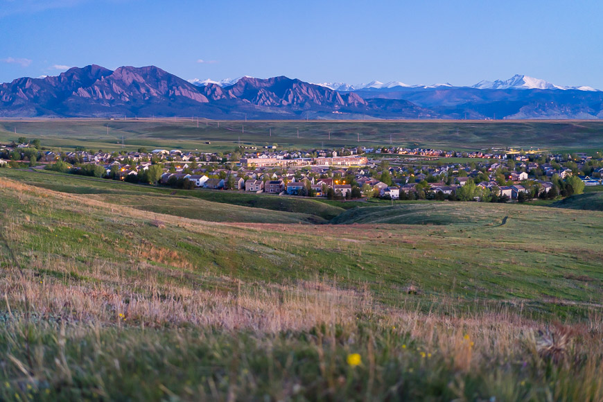 A small town is nestled in a green valley with the Rocky Mountains in the background under a blue sky.