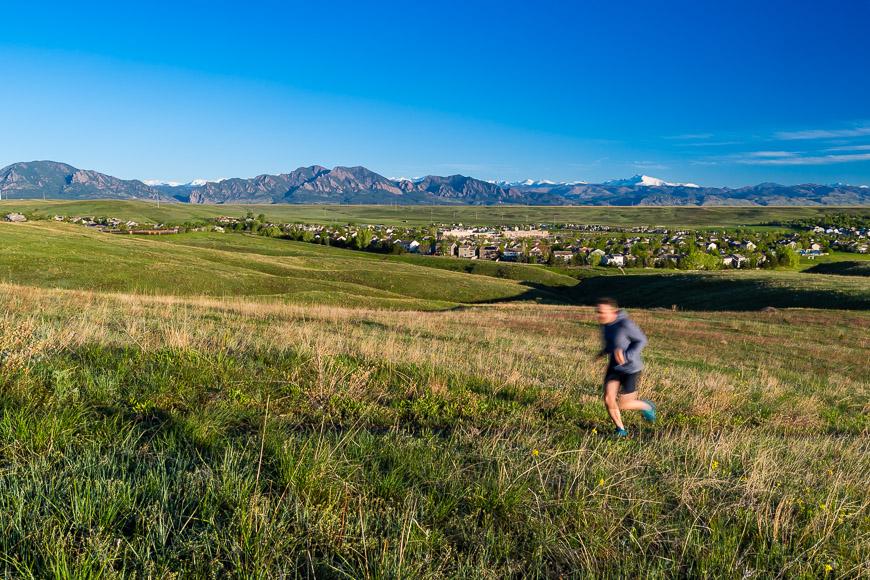 A person runs across a grassy field with a distant view of a town and mountain range under a clear blue sky.