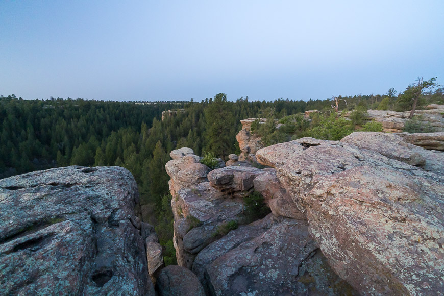 Rocky cliff edge overlooking a dense forest under a clear sky.