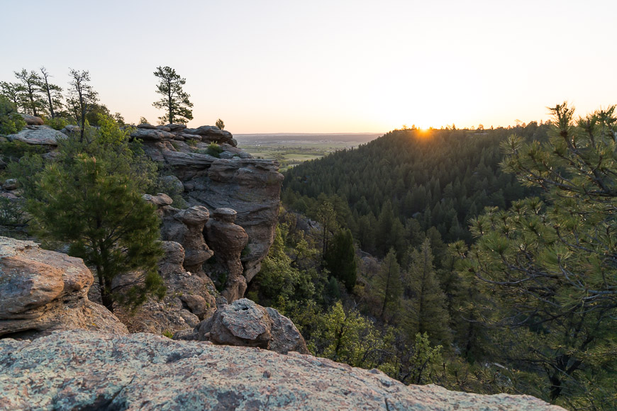 A rocky cliff overlooks a forested valley at sunrise, with trees and distant landscapes visible under a clear sky.