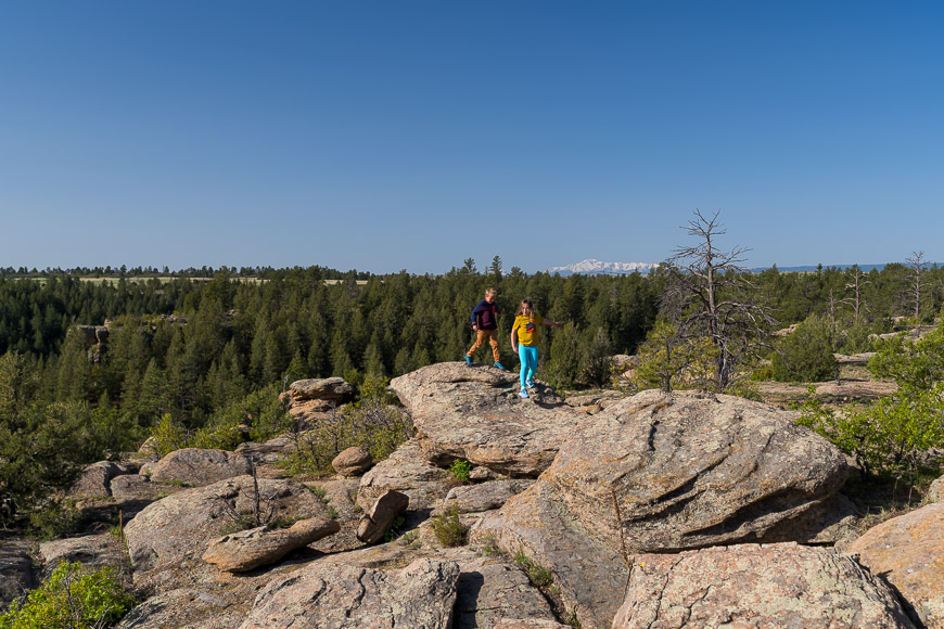 Two people walking on large rock formations surrounded by a forested landscape under a clear blue sky. Snow-capped mountains are visible in the distance.