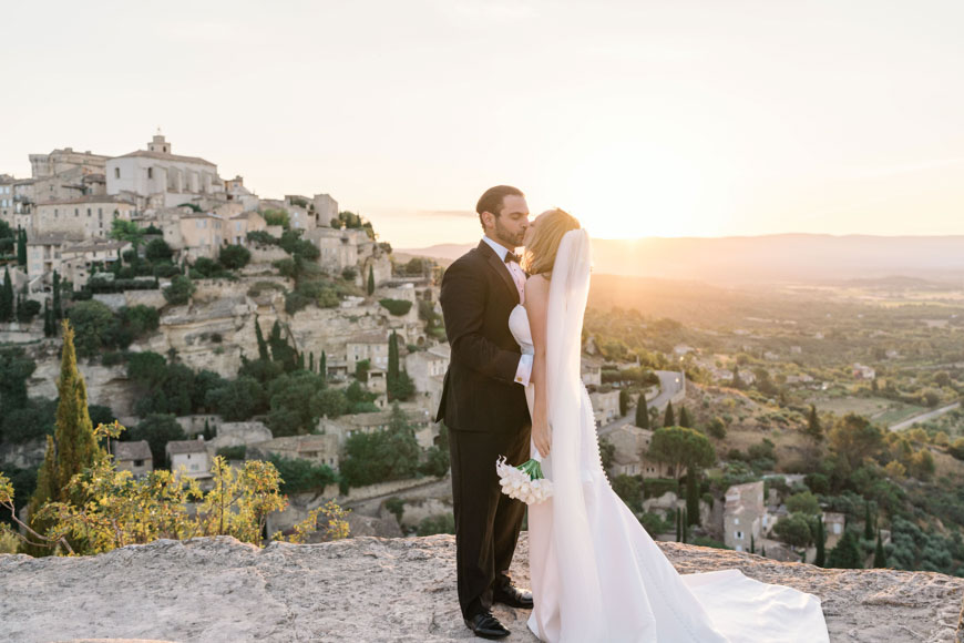 A bride and groom stand on a rocky ledge, kissing at sunset, with a historic hillside village in the background.