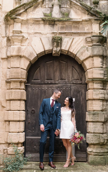 A couple in formal attire stand holding hands in front of an arched stone doorway. The person on the left wears a suit, and the person on the right is in a white dress holding flowers.