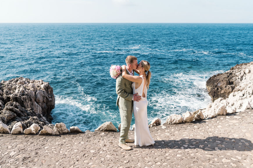 A couple embraces and kisses on a rocky seashore with the ocean in the background.