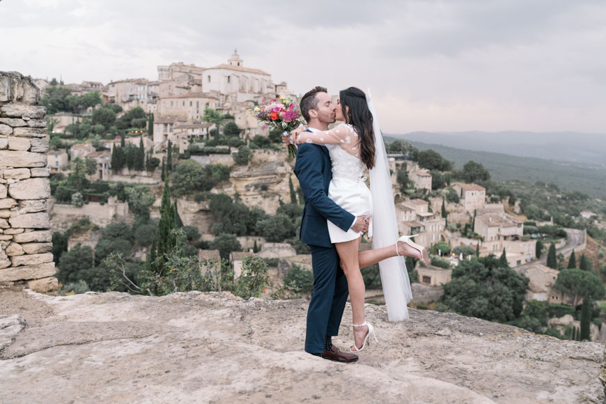 A couple in wedding attire embraces on a rocky ledge with a historic European village in the background.