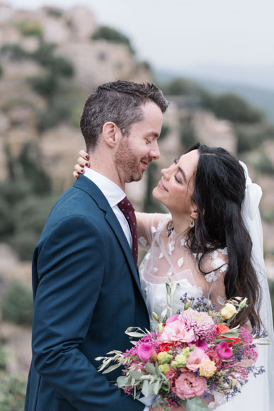 A couple embracing outdoors, with the woman holding a colorful bouquet and the man wearing a dark suit. The background features blurred greenery and stone structures.
