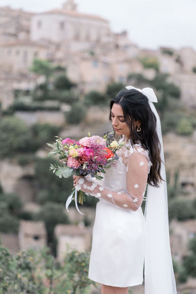 A bride in a white dress holds a colorful bouquet while standing outdoors with a scenic, blurred background.