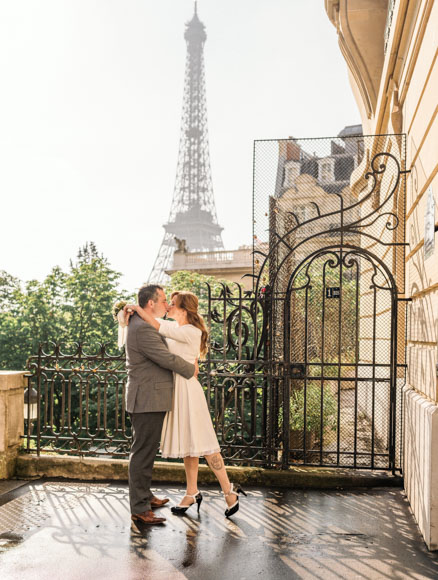 A couple embraces near a decorative gate with the Eiffel Tower in the background on a clear day.