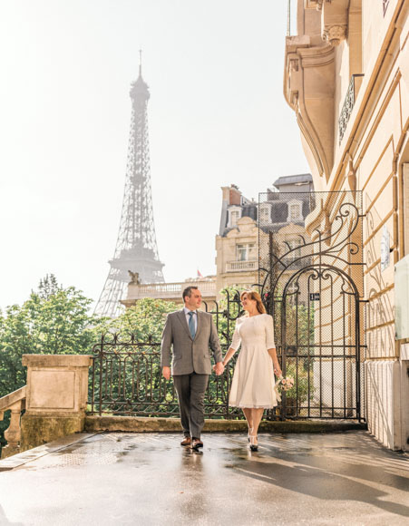 A couple holding hands walks along a Parisian street with the Eiffel Tower in the background.