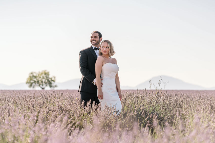 A couple stands in a lavender field under clear skies. The woman wears a white dress, and the man wears a black suit. A tree and mountain are visible in the background.