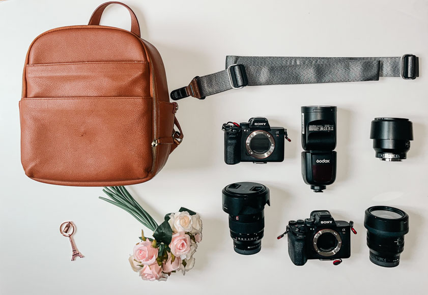 Brown backpack, camera gear, flowers, and a keychain on a white surface.