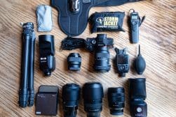 Overhead view of a photography equipment set including a camera, lenses, tripod, cables, remote shutter release, camera covers, flash, blower, and filters arranged neatly on a wooden surface.