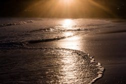A serene beach scene with gentle waves washing onto the shore at sunset. The sun is low on the horizon, casting a golden reflection on the wet sand and water.