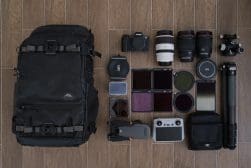 A collection of photography gear laid out on a wooden floor, including a large backpack, camera, lenses, drone, filters, tripod, remote control, and cases.