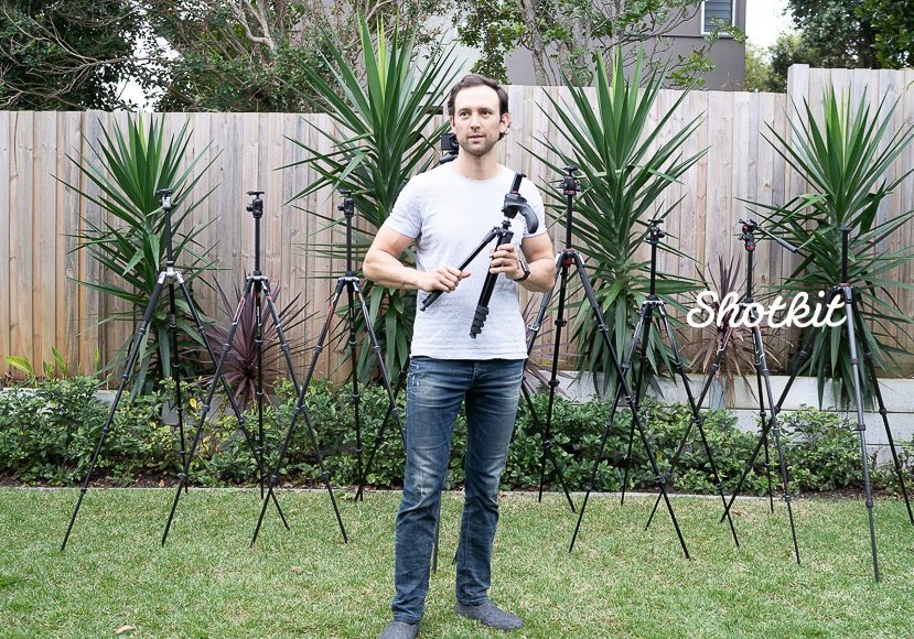 A man stands in a garden holding a camera tripod, surrounded by various tripods of different sizes, with plants in the background. A man stands in a garden holding a camera tripod, surrounded by various tripods of different sizes, with plants in the background.