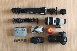 Camera gear layout, including two tripods, a drone, a camera with lens, three additional lenses, a camera controller, memory cards, and an apple, all placed on a wooden surface.