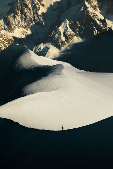 A lone figure stands on a snow-covered mountain slope with jagged peaks in the background.