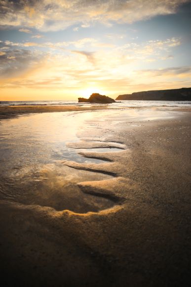 Sunset view on a beach with rippled sand patterns leading to the water. The sky is partly cloudy, and the horizon is highlighted by the evening light.