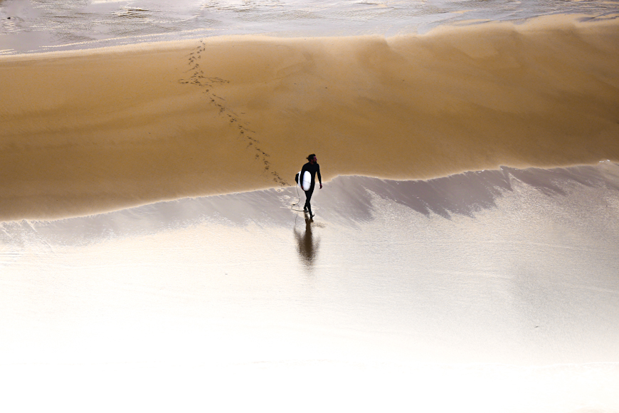 A person with a surfboard walks along a sandy beach with footsteps visible in the sand.