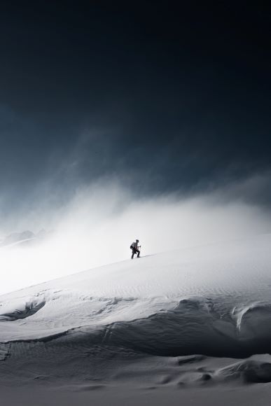 A lone hiker walks up a snowy incline under a dark, cloudy sky, surrounded by mist.