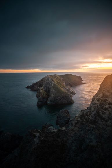 A rugged coastline with rocky cliffs overlooks the ocean at sunset, with a dramatic sky casting shadows on the water.