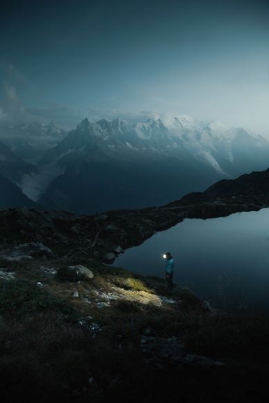 Person with a flashlight standing by a calm lake in a mountainous landscape during twilight.