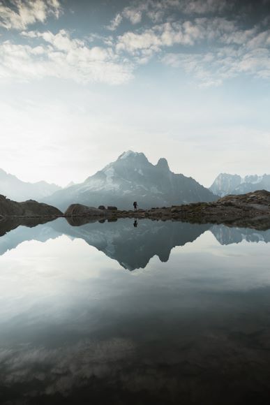 A lone person stands by a calm mountain lake with a clear reflection of snowy peaks and scattered clouds above.