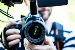 Person holding a DSLR camera with a large lens, facing the viewer. Background is blurred and person is wearing a striped shirt.