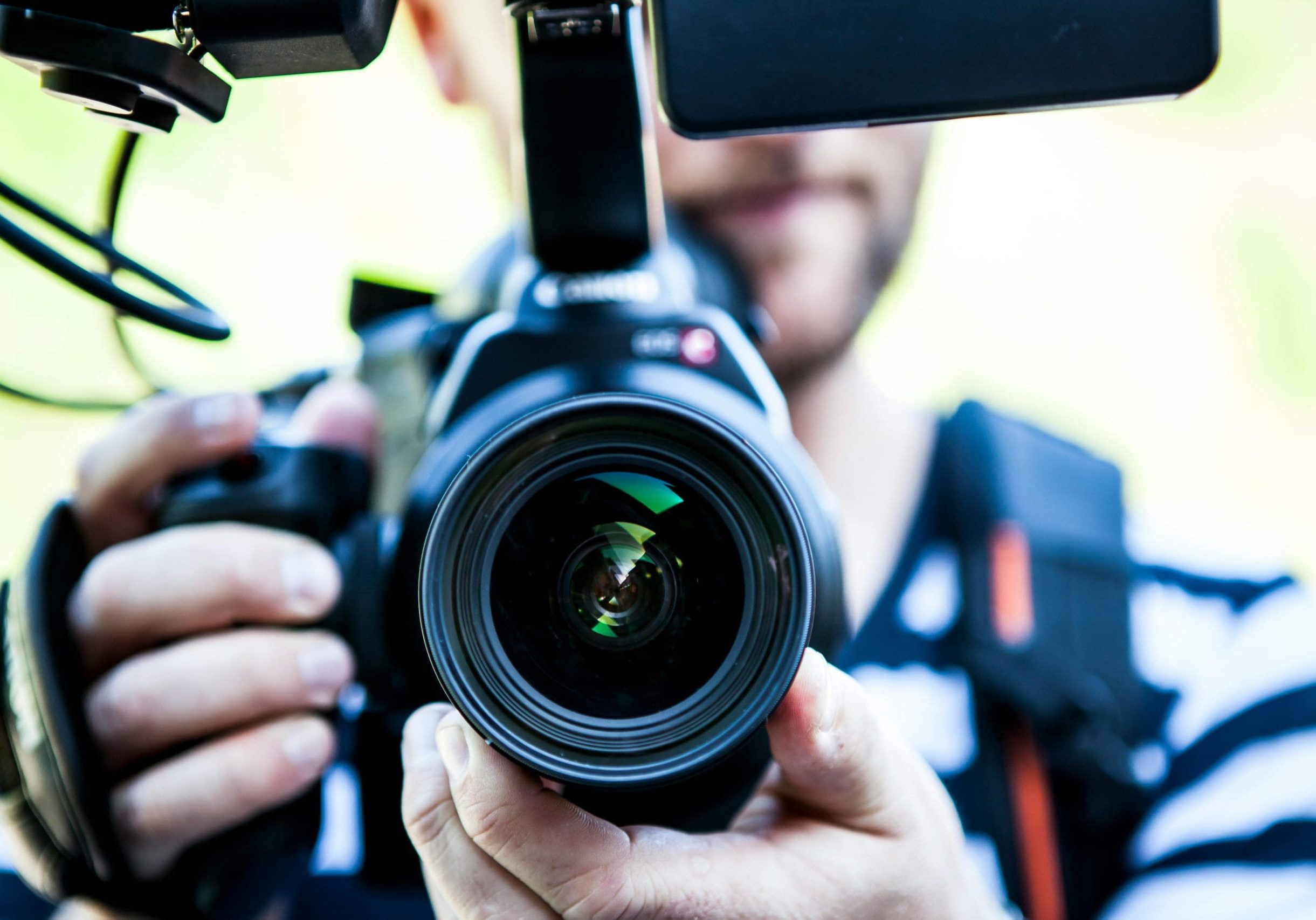 Person holding a DSLR camera with a large lens, facing the viewer. Background is blurred and person is wearing a striped shirt. Person holding a DSLR camera with a large lens, facing the viewer. Background is blurred and person is wearing a striped shirt.