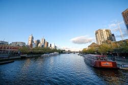A river with a boat in the foreground flows through a city with tall buildings and a bridge in the background under a clear blue sky.