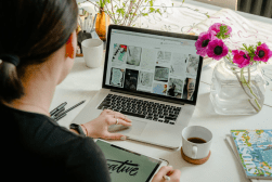A person is working at a desk with a laptop displaying art images, a tablet, a cup of coffee, and flowers in a vase nearby.