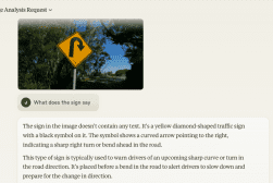 A yellow diamond-shaped traffic sign with a black arrow indicating a sharp right turn or bend ahead. The sign is placed beside a road with trees in the background.