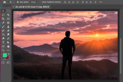 Person stands on a hilltop overlooking a sunset with clouds and mountains, as seen on a computer screen with photo editing tools.