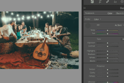 Group of people enjoying a meal outdoors at night under string lights, with a table full of dishes and a guitar nearby.