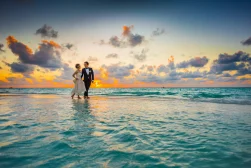 A bride and groom walk on a beach at sunset, with the ocean waves and a cloudy sky in the background.