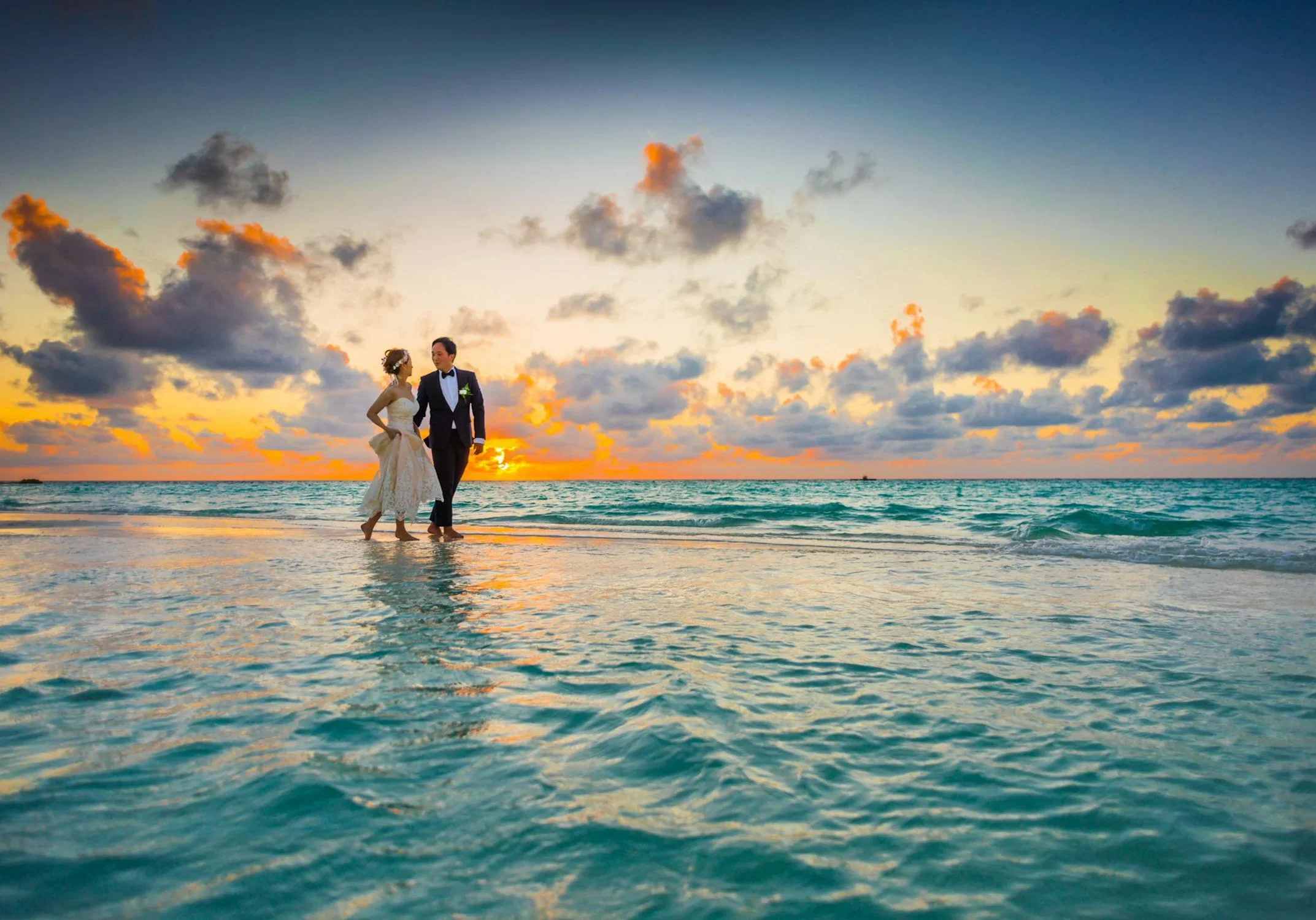 A bride and groom walk on a beach at sunset, with the ocean waves and a cloudy sky in the background. A bride and groom walk on a beach at sunset, with the ocean waves and a cloudy sky in the background.