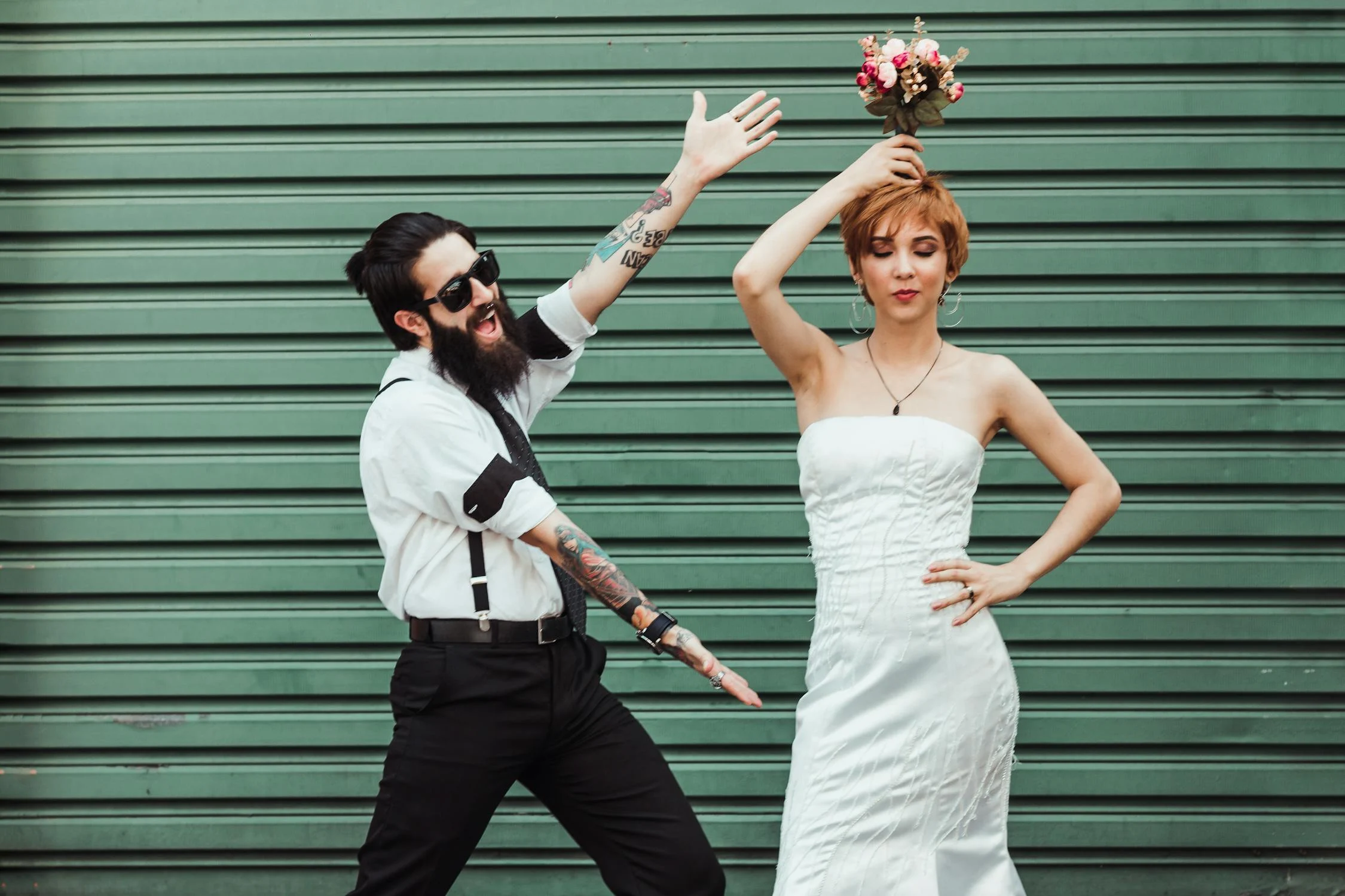 A man with a beard and tattoos gestures dramatically towards a woman in a white dress who holds a bouquet above her head, both standing in front of a green striped background.