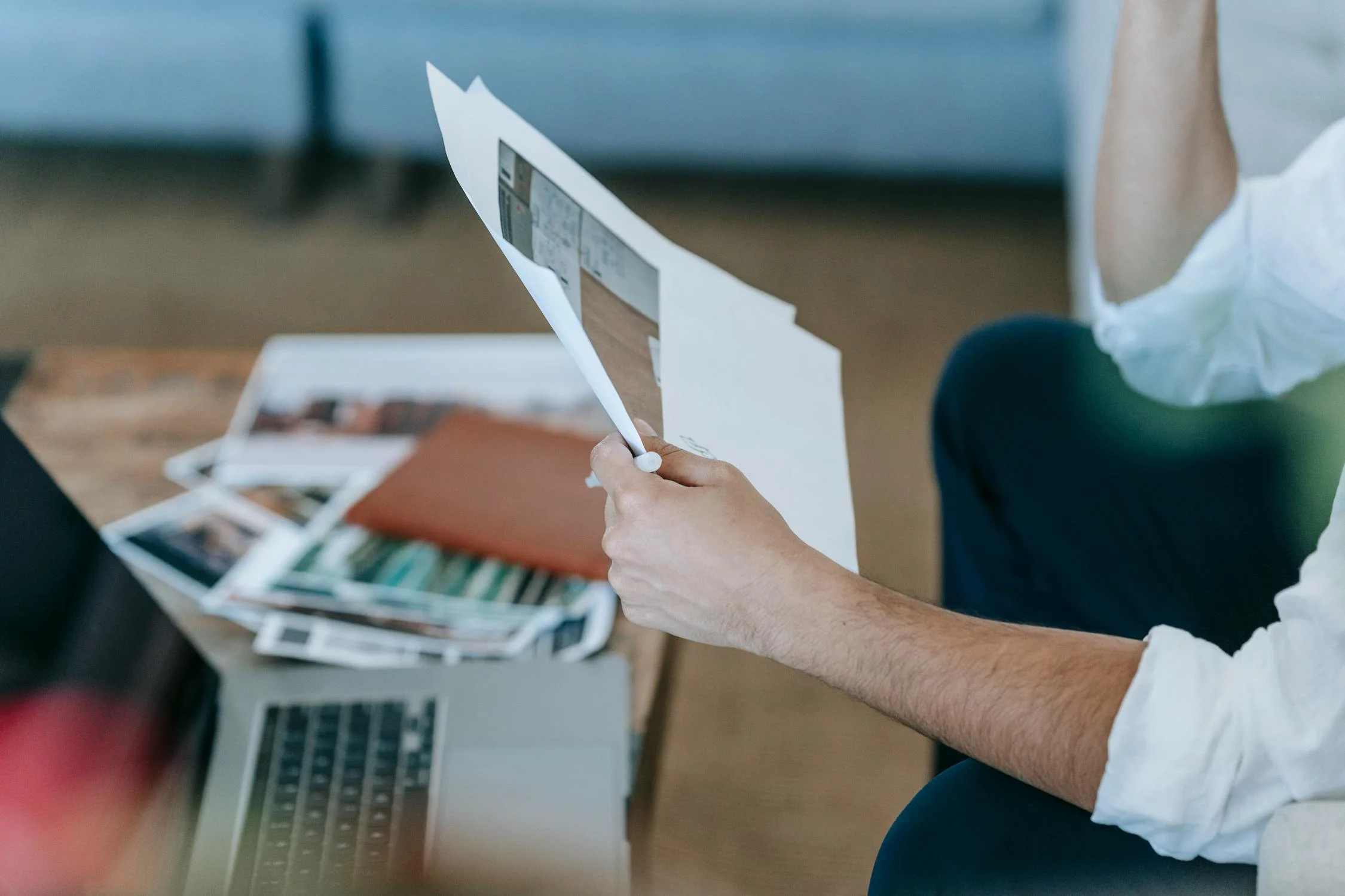Person holding and reading documents next to a laptop and a pile of papers on a wooden table.