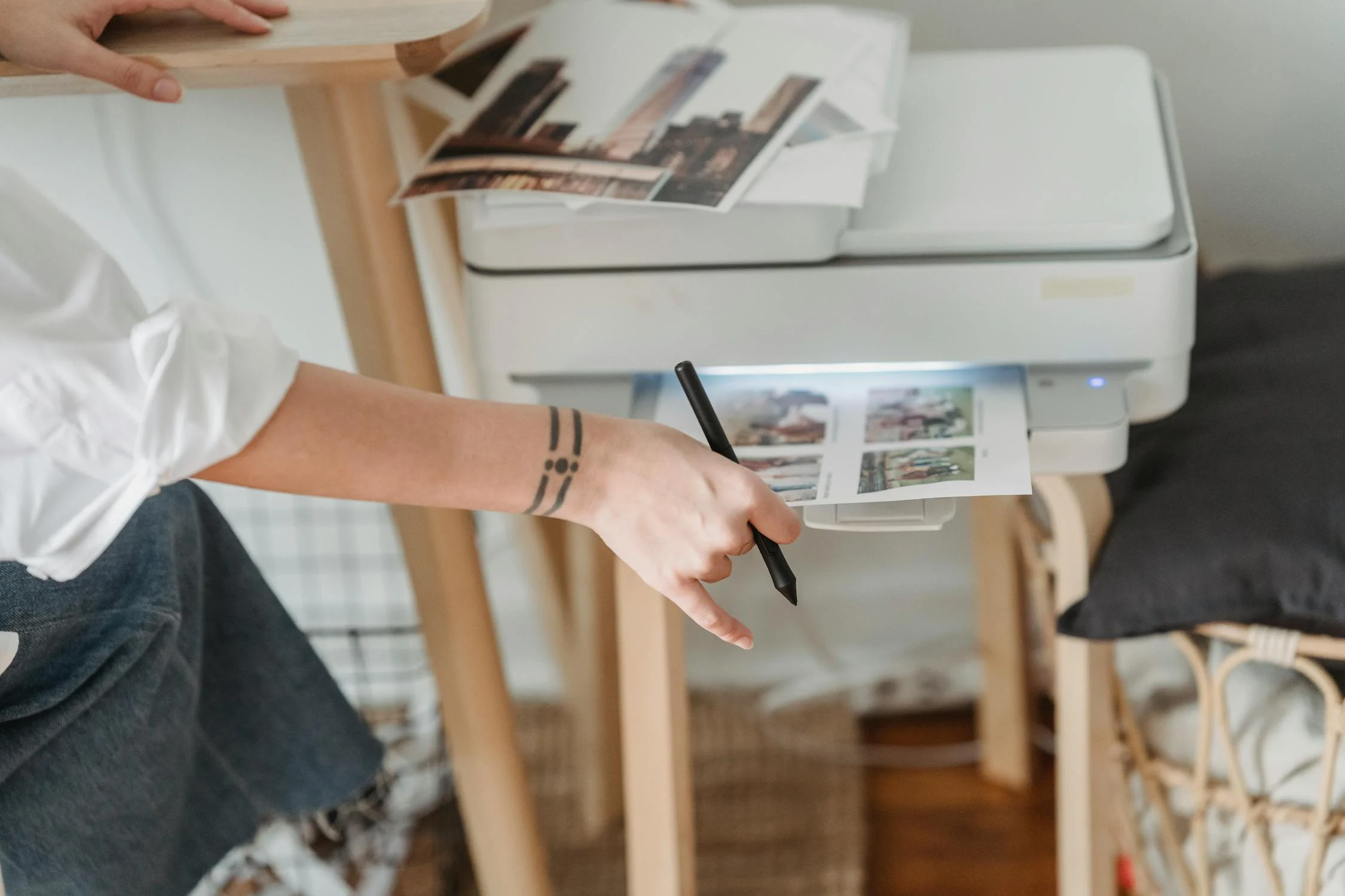 A person uses a stylus pen to edit images on a printer. The printer and photos are on a wooden table.